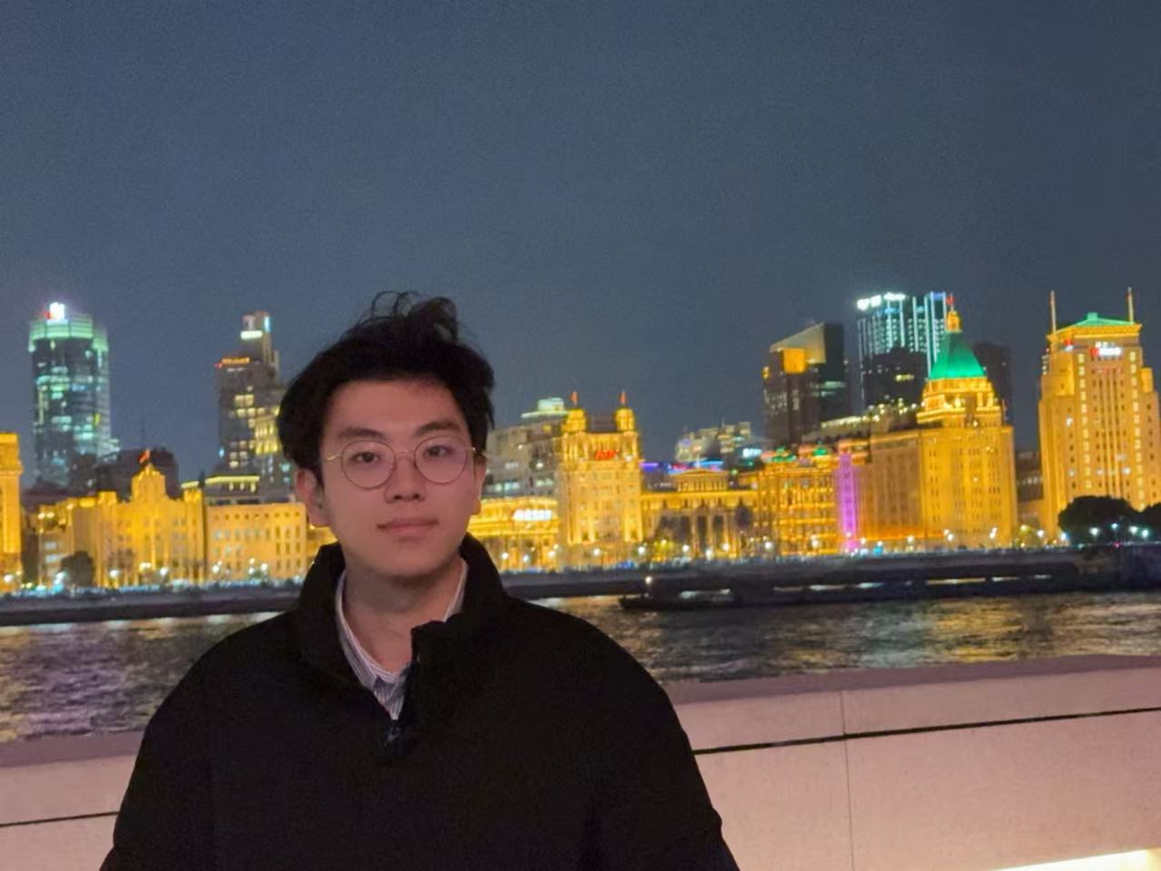 Yehang Zhang by the river at night, with illuminated buildings behind him